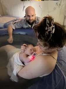 Mother and newborn in water birth tub, partner looking on with joy. 
